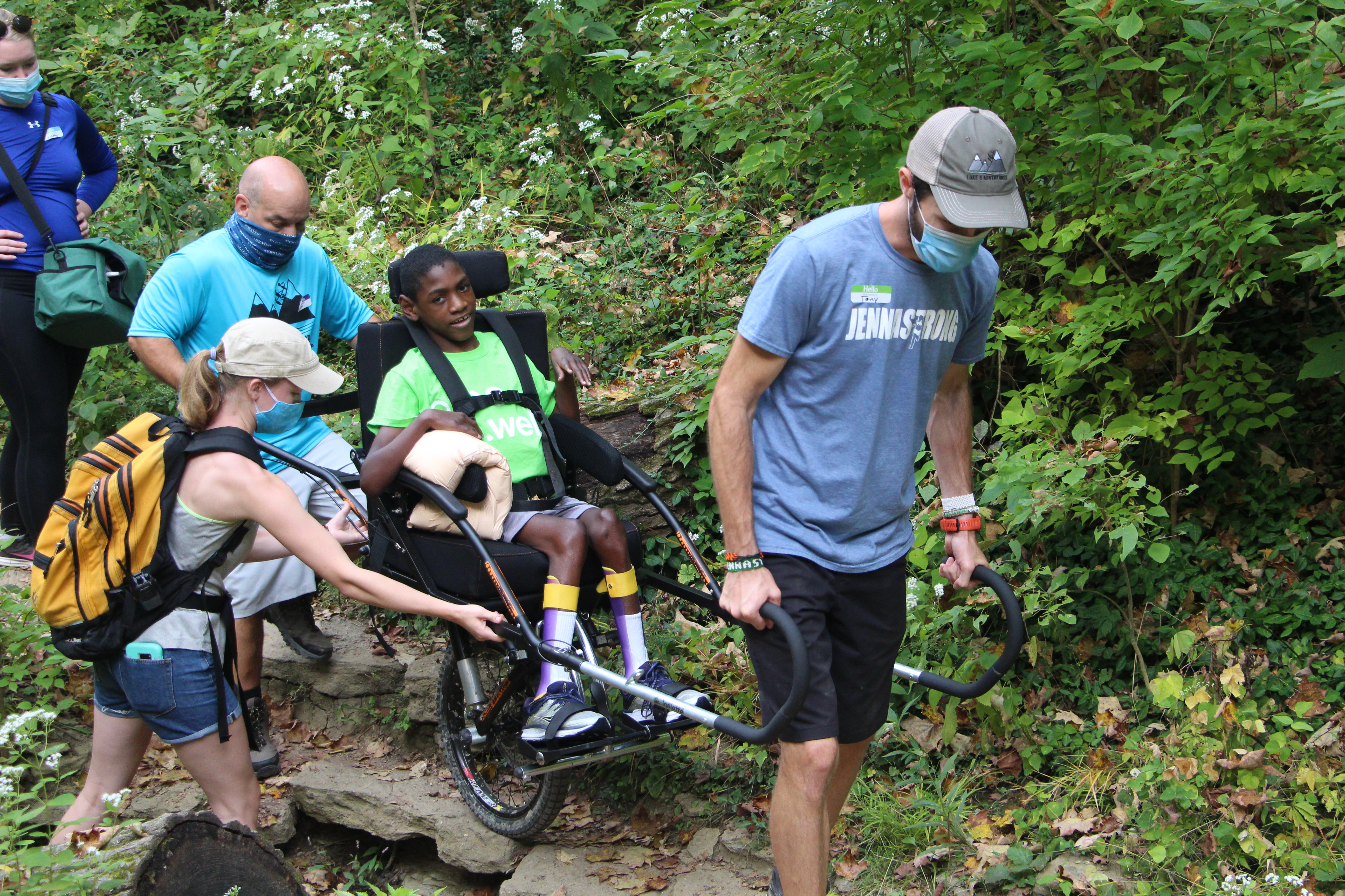 Child in an all-terrain wheelchair crossing over a rocky terrain with the assistance of 3 adults.